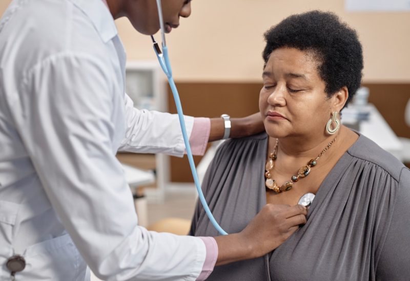 Medium close up shot of mature Black woman patient sitting with closed eyes in clinic while doctor conducting auscultation