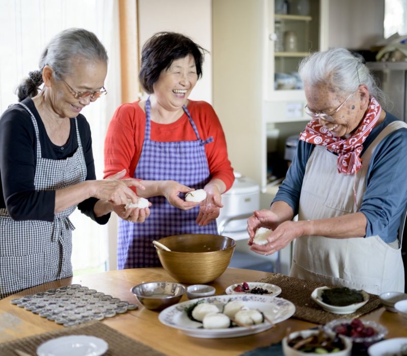Three older women standing round a table in a kitchen, making sushi.